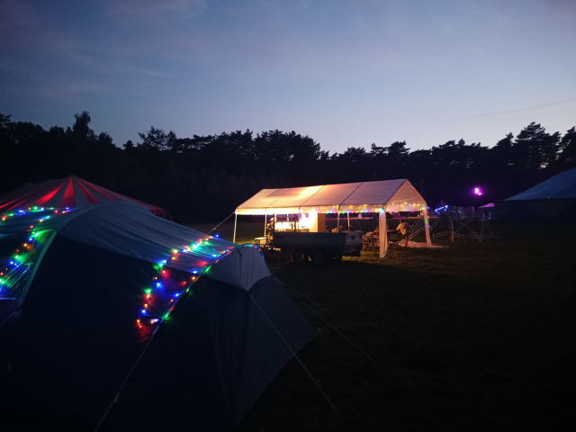 Tents with colourfull lights. The sky is still lit as it is dusk, but the trees at the background are dark