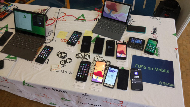 A table with the famous Linux Mobile tablecloth, a bunch of phones running Linux Mobile with various UIs, a laptop with GNOME and a tablet with keyboard with Plasma