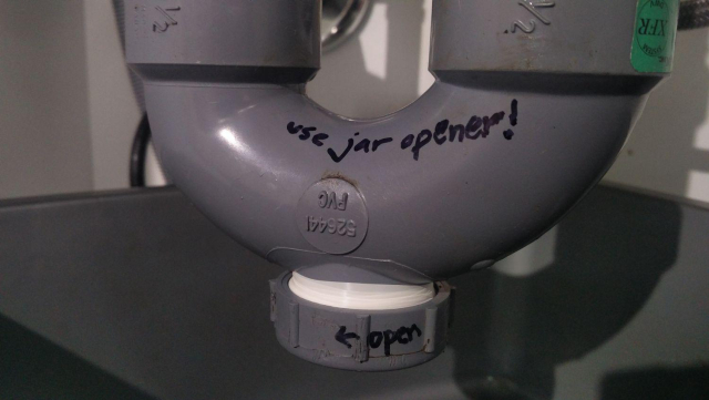 A P-trap bend pipe under a sink, annotated with a sharpie to say:

"Use a jar opener!" (written on the pipe)

"← open" (and "close →") on the cap to indicate the rotation direction for unscrewing and screwing the cleanout cap