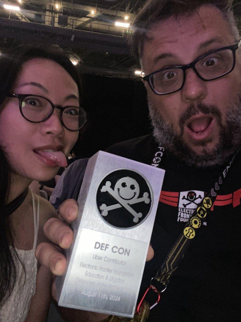 A woman with glasses and her tongue out next to a man with a beard and glasses looking surprised. He is holding a silver rectangle award with the DEFCON logo and the the phrase Uber Contributor Electronic Frontier Foundation. 
