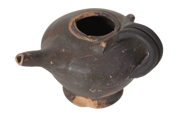 A photo of a clay pot against a plain white background. The pot is a greyish-brown color, with hints of terra cotta where the paint has chipped, revealing the ceramic material it is made of. 

The pot is chipped and flaked, suggesting considerable age. It has a small handle, a circular opening on the top, and a small round base.

Notably, it has a tiny spout, much narrower than would be usual for a pot this size—perhaps just the right size for an infant with a cleft palate to drink from it.