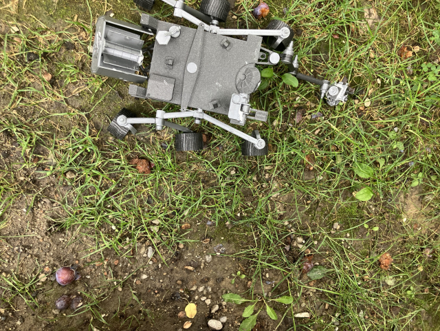 This image shows a 3D printed model of the Mars Perseverance Rover placed on the ground. The ground is grassy and a bit muddy with some visible pebbles and small plants.  Overall Visual Description of the Rover: The rover model is primarily gray and appears to have a detailed structure resembling the actual Mars Perseverance Rover. It is a six-wheeled vehicle with a box-shaped body and several protruding instruments and appendages. 1. Main Body: - Rectangular Box Structure: This is the central part of the rover which contains the main components and instruments. It is roughly squarish and houses the essential systems, including telecommunications and power. 2. Mast (Central Post): 