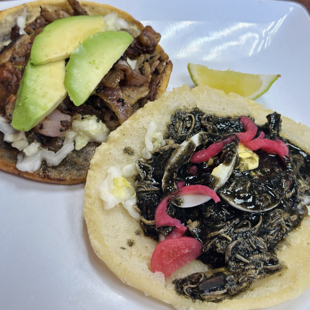 A photo of a panucho and salbut topped with various meats. The salbut has shredded chicken that looks black, as it comes with the soup / sauce from the delicious relleno negro that they make 