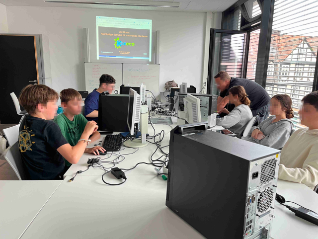 A group of students are shown installing GNU/Linux on discarded computers. The students have just learned about how software can be responsible for environmental harm. In the background the slides used in the presentation, including the KDE Eco logo, are visible. 