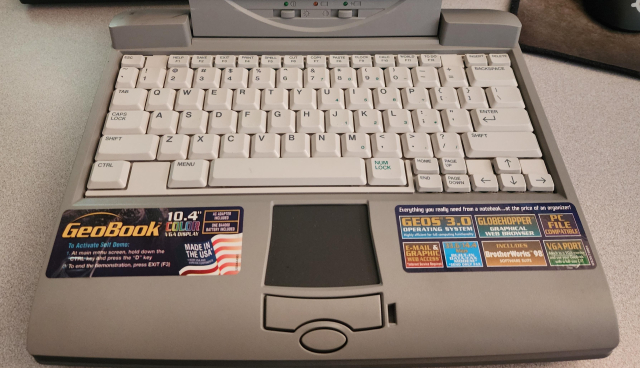 The inside of a grey 90s laptop. It's got a touch pad, light grey keyboard, and two big stickers.
The left calls it a GeoBook 10.4" Color VGA Display, made in the USA, and says how to activate a "demo".
The right side says "Everything you really need from a notebook... at the price of an organizer!" with smaller labels for Geos 3.0, Globehopper Graphical Web Browser, PC File Compatible, Email & Graphic Web access, 33.6/14.4 modem, includes BrotherWorks '98, and VGA port 