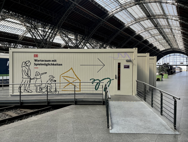 Ein Wartebereich für Kinder aus Schiffscontainern mit verspielten Linienzeichnungen einer Familie und eines spielenden Kindes. Der Container befindet sich in einem geräumigen Bahnhof mit einer hohen, gewölbten Decke und sichtbaren Bahngleisen im Hintergrund.