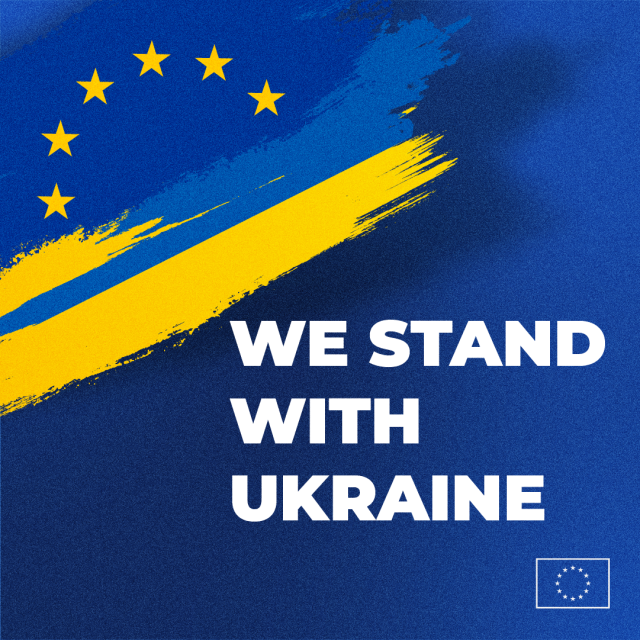 The image shows a design featuring a blue background with a diagonal yellow brush stroke, representing the Ukrainian flag. The European Union stars are depicted in a semicircle at the top left corner of the image, integrated into the brush stroke. The text "WE STAND WITH UKRAINE" is boldly written in white across the lower half of the image. A small European Union flag is placed at the bottom right corner. 