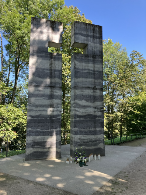 Zwei steinerne Blöcke, die mit ihrem Abstand ein Kreuz bilden vor dem Gräberhang im Wald, darunter ein Blumenkranz mit Schleife und brennende Kerzen