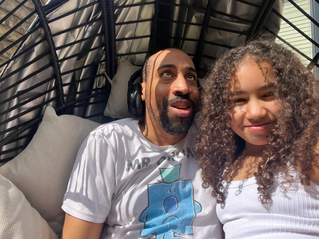 The image shows a man and a young girl sitting closely together in a cozy, outdoor setting. The man has a shaved head, a beard, and is wearing headphones around his neck. He is dressed in a white T-shirt featuring a cartoon character with the words "Mr. Grumpy" on it. The young girl has long, curly hair and is wearing a white tank top. They are sitting in a hanging chair with a woven structure, and there are light-colored cushions behind them. Sunlight is streaming in, casting shadows and creating a warm, relaxed atmosphere. The man is looking upwards with a smile, while the girl is smiling directly at the camera.