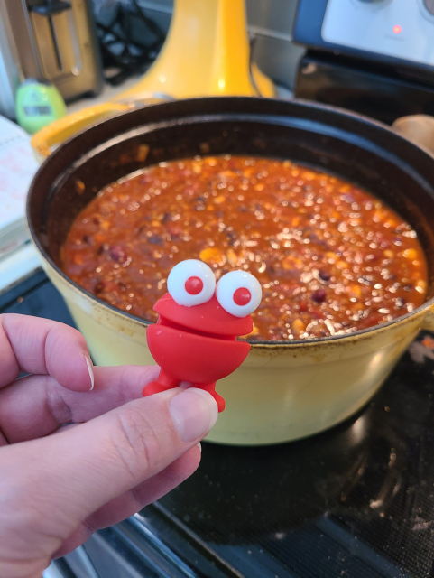 Picture of someone's hand holding a small red googly-eyed monster. The silicone monster is essentially an ellipse with a very wide mouth nearly bifurcating its entire body, with feet.
A very large Dutch Oven full of fresh unsimmered chili lurks on the stove in the background.