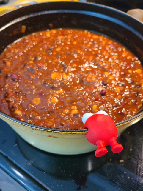 Small red elliptical silicone monster, face down and feet towards the camera, biting the edge of a Dutch Oven full of chili with its entire body.