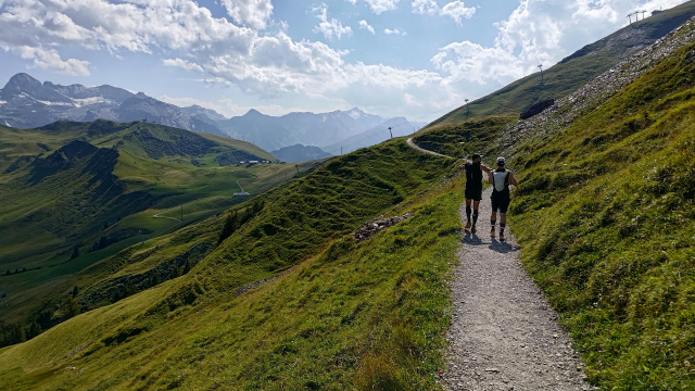 Two hikers walk along a narrow gravel trail through green, rolling hills. Majestic mountains with snow-capped peaks rise in the background under a partly cloudy sky. The trail winds upward, and ski lift poles are visible, hinting at winter activities. The scene is peaceful, with soft sunlight enhancing the natural beauty.