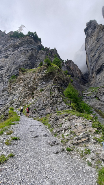 Two hikers are making their way up a steep, rocky trail that winds through a dramatic mountain pass. Towering cliffs rise sharply on either side, with patches of greenery clinging to the rugged terrain. The sky is overcast, with clouds hanging low, adding to the sense of height and isolation. The scene is rugged and intense, highlighting the challenging nature of the hike.