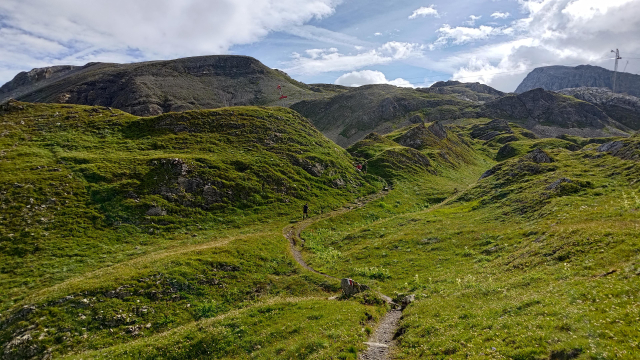 A narrow dirt trail winds through green hills, leading toward rugged, rocky mountains in the background. The landscape is lush, with patches of grass covering the terrain. A few hikers can be seen in the distance, walking along the path under a partly cloudy sky. The scene is serene, capturing the raw beauty of a mountainous area.