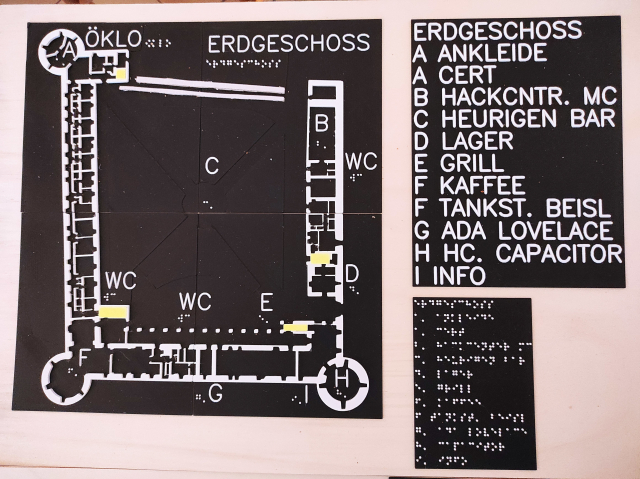 Taktiler Plan vom Håck ma's Castle. Erdgeschossgrundriss des Schlosses in Ottenschlag mit vier runden Ecktürmen, West-, Süd- und Osttrakt. Orte am Plan sind mit Buchstaben in taktiler Profilschrift und Brailleschrift markiert. Eine Legende in Profil- und Brailleschrift erklärt die Bedeutung der Buchstaben am Plan. Weiße Zeichnung und Schrift sowie gelb markierte Räume auf schwarzem Grund auf einer Holzplatte.