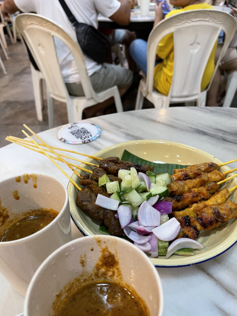 A plate of satay, a Malaysian/Indonesian dish featuring roasted meat skewers, including both chicken and mutton options. It's served with a spicy peanut sauce, along with sliced raw onions and cucumbers.