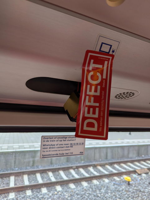 A red sticker saying DEFECT covering electrical outlet on a train with something hanging out of it.