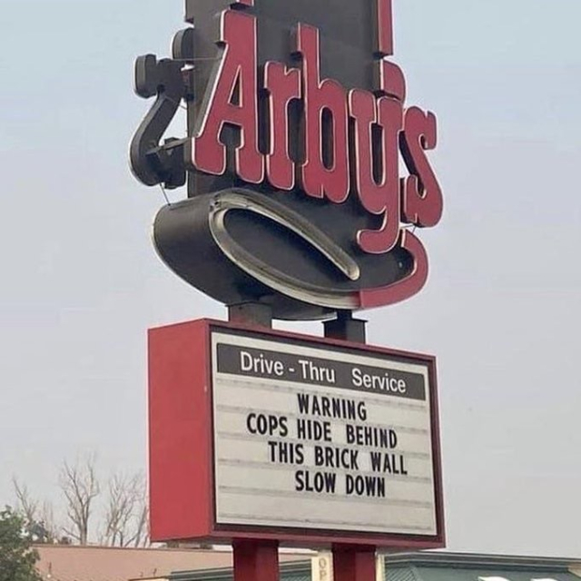 A sign outside an Arby's restaurant warns drivers that police hide behind a nearby brick wall and advises them to slow down.