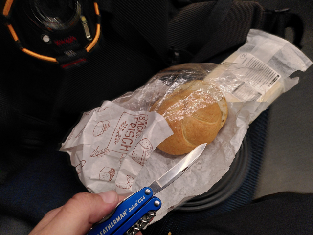 A blue leatheman juice CS4, a kaiserbrochen, and a block of cheese. On a plate on a train seat.