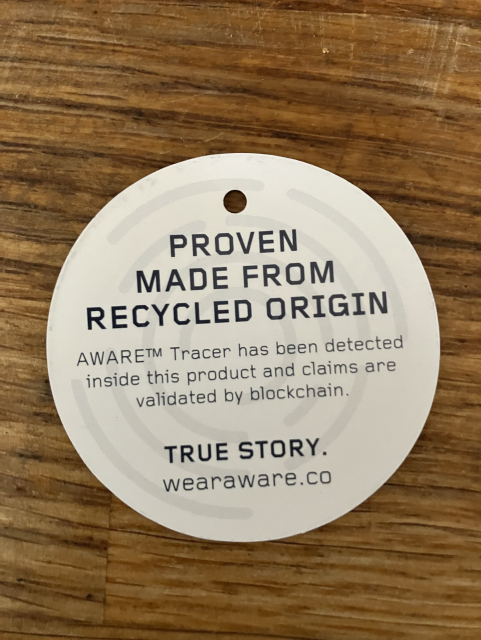 A circular tag from a tote bag. It lies on a wooden surface stating it is "PROVEN MADE FROM RECYCLED ORIGIN." It mentions the AWARE™ Tracer for validation of claims via blockchain. The tag includes the text "TRUE STORY" and the website wearaware