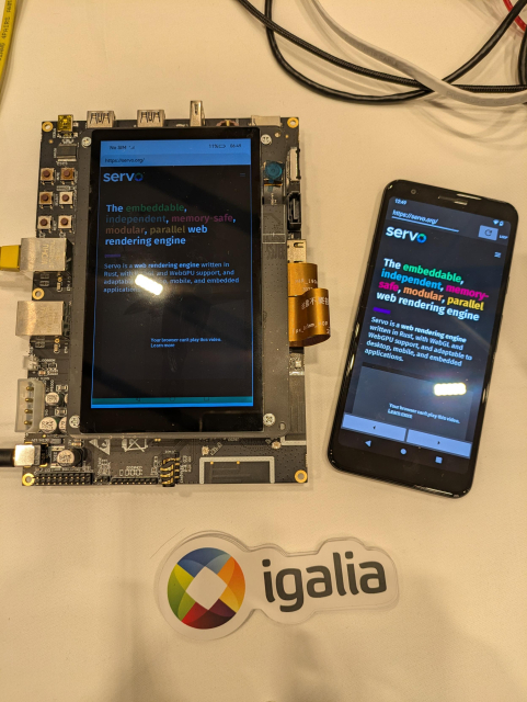 Picture of a Rockchip board and an Android phone, both running Servo and showing the servo.org homepage. At the bottom of the image there are some stickers with Igalia logo.