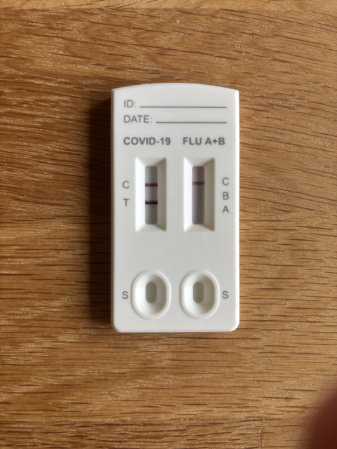 A positive COVID-19 and flu A+B rapid test kit displayed on a wooden surface. The test shows two result windows labeled for COVID-19 (C and T) and flu (A and B), with areas for sample collection indicated as "S."