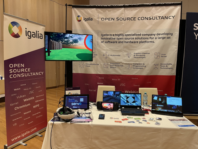 Igalia Booth B8 at OSSSummit EU 2024 including some Igalia banners and a table with multiple computers running demos of Wolvic, Servo, WPE WebKit and graphics. Among them, a Steam Deck and some VR glasses.