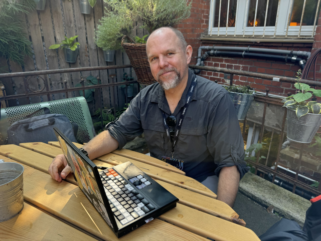 A picture of Mike Hoye working from his MNT Reform laptop from a wooden table in the garden of the Matrix Conference venue