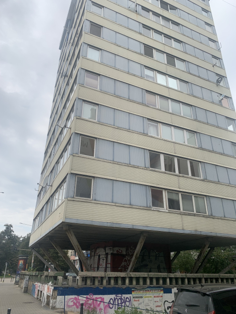 A tall, modern building elevated on stilts, featuring a façade of yellow and gray panels. The structure has numerous windows and is adorned with graffiti at the base.