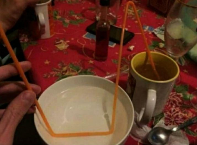 A straw leads of of a mug of tea on a table with an ornate red tableclothe. The straw bends and passes through a bowl of water, presumably water cooling it before bending up so the tea can be drunk at mouth level