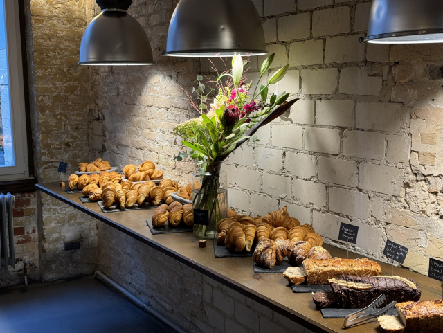 A picture of a wooden shelf with fresh pastries under a warm light. There are flowers between the pastries.