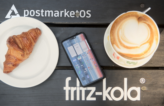 nearly straight down, high angle shot of a dark brown to black wooden plank outdoor table. On it are a croissant on a china plate to the left, and a impressively large coffee with milk to the right, between them is a OnePlus 6 running postmarketOS with multiple apps open (Foliate, Railway, Meteocool). The table has a fritz-kola branding on the bottom left and a postmarketOS logo in the top left. The postmarketOS logo might look convincing unless zoomed in extremely closely, because there is suspiciously little chromatic apparition, showing it was added later, and wasn't there when the photo was taken