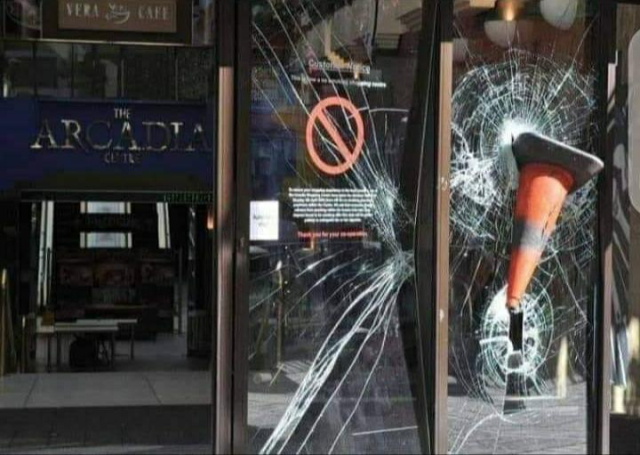 A photo of two floor to ceiling shop windows. One is badly cracked having been hit by something. The second is smashed even worse and has an orange traffic cone embedded in the window