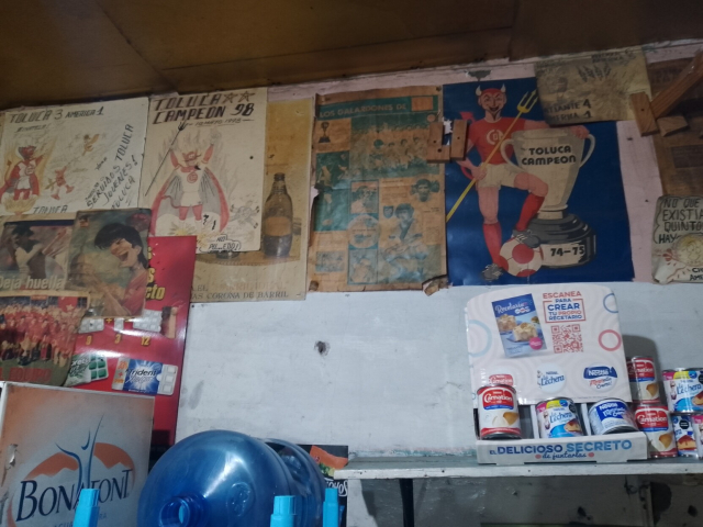 Pared atiborrada de afiches de equipos de futbol de diferentes épocas 70's - 90's. Estás hojas venían en la sección deportiva de los periódicos dominicales. Por debajo de los afiches se observa un refrigerador de Bonafont, productos Nestlé y un garrafón azul acostado.