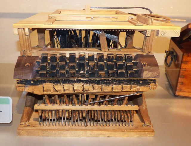 Prototyp einer Schreibmaschine, Peter Mitterhofer, 1864; Exponat im Technischen Museum Wien. Eine Schreibmaschine aus Holz und wenigen Metallteilen. Ohne Verkleidung sind zahlreiche mechanischen Hebel und Details erkennbar. Die schwarze Farbe an den Tasten aus Holz spiltert ab.