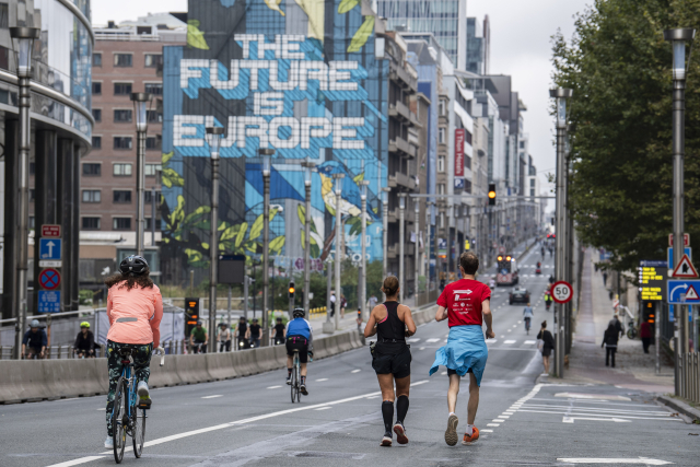 This image shows a street scene with cyclists and runners moving towards a large mural in the background that reads "THE FUTURE IS EUROPE." In the foreground, a person on a bike wearing a pink jacket and helmet is on the left, while two runners are on the right, one wearing a red shirt and the other in black running gear. Additional cyclists are seen further down the road, heading toward the same direction. The surrounding area features tall buildings and urban infrastructure, with street signs, lampposts, and some pedestrian activity. The atmosphere is active, with the mural being a prominent focal point. 