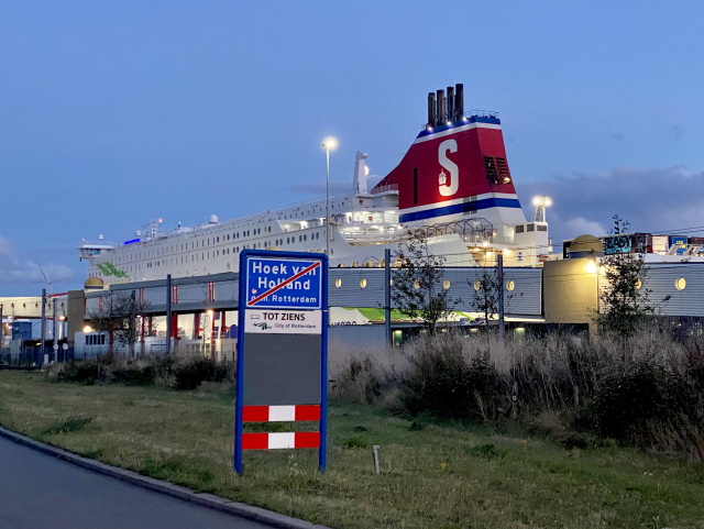 Evening. A blue road sign with “Hoek van Holland” struck out with a red bar. Beyond is a white ship with a red and blue chimney and a bug white S.