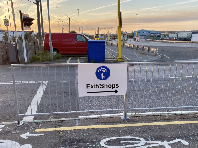 A sign on a railing protecting the footpath from the road reading “Exit/Shops” and pointing to the right.