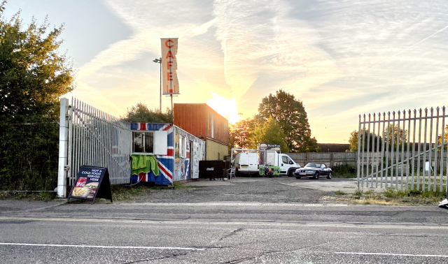 An industrial estate with a big iron fence. On it are a bunch of shipping containers and vans cosplaying as a cafe and chippie.