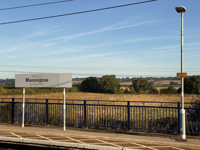 View across the tracks to a platform with a big white station sign saying “Welcome to Manningtree for Dedham Vale” and a lamp. Beyond a black railing are fields and trees and rolling hills.