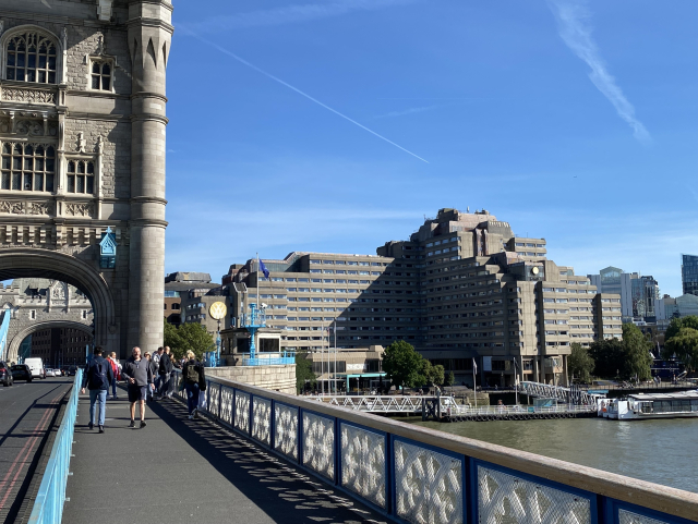 Walking across the Tower Bridge. You can see half of one of the bridge towers on the left. The focus is on a large grey concrete building on the right.