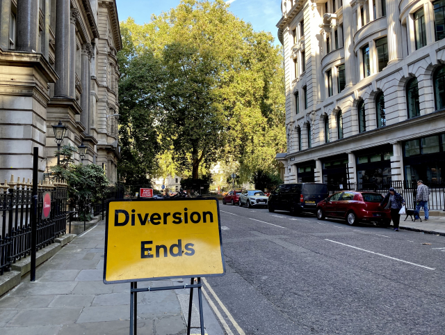 A yellow street sign reading “Diversion ends” stands in the sidewalk of a city street with tall
buildings and trees off in the distance.