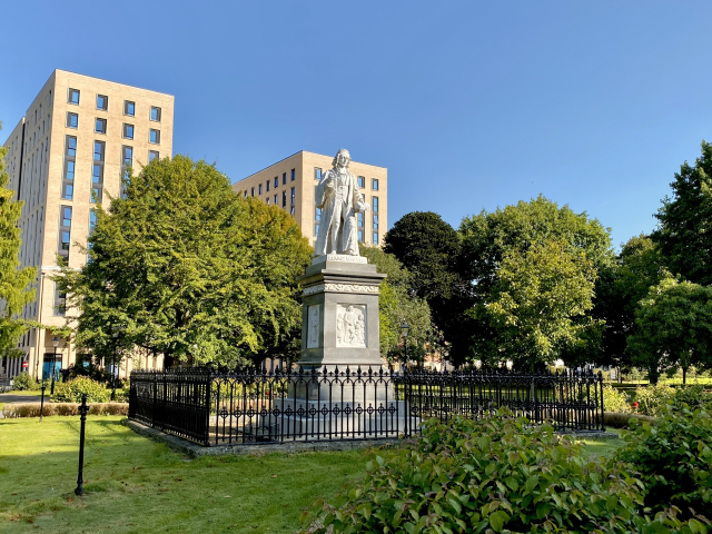 A statue of a man in a wig on a plinth in a park. Tall buildings beyond, blue skies.