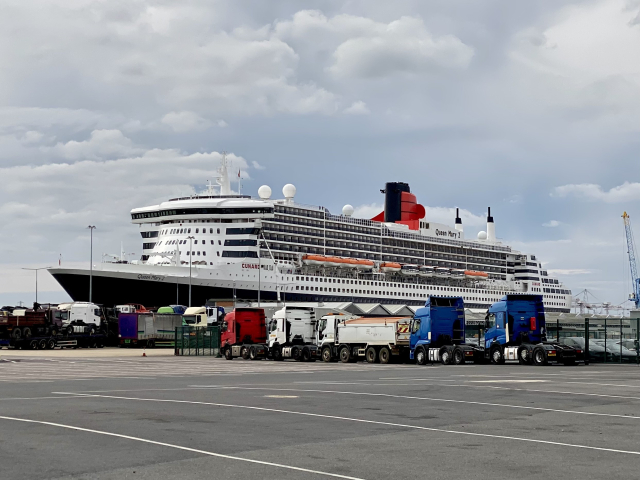 Shot across a harbour peer with slender black and white ship beyond claiming to be Queen Mary 2.