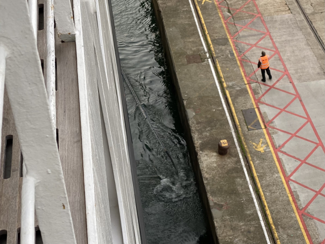 View down the side of a ship. A dock worker has just cast the a line into the water. 