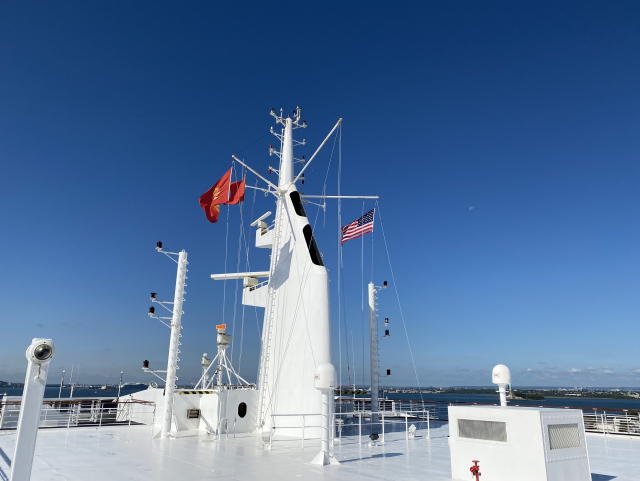 The white forward mast of a ship. The red Cunard flags is flying port side together
with a red ensign and the US flag on starboard.