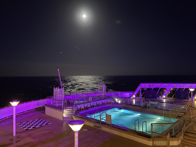 A full moon hangs over the aft pool deck lit in purple and the see beyond.