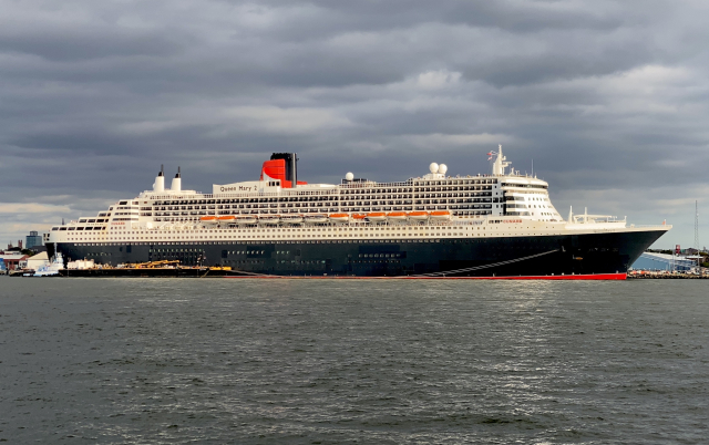 Side view of a long slender passenger ship, red bottom, black hull, white superstructure, and a red funnel. Next to the funnel it says Queen Mary 2 in large Gill letters.