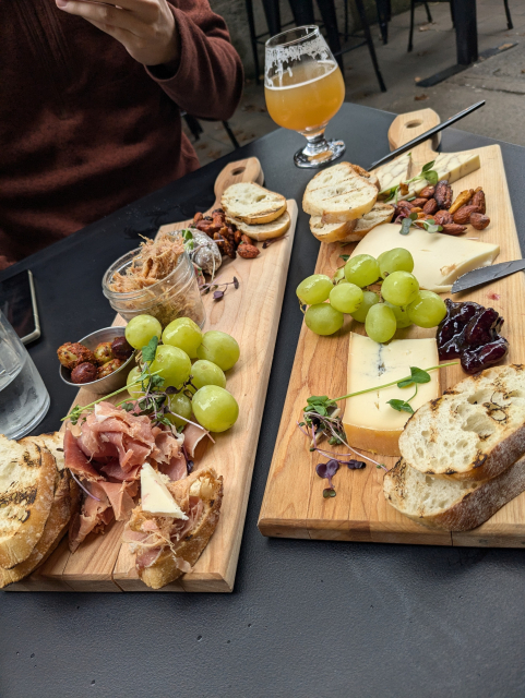 A board of charcuterie (prosciutto, dry sausage, duck Rillette, nuts, grapes, bread) and a board of cheese (blue, mild and nuts) with jams and chutneys. There is a glass of milkshake IPA at the back