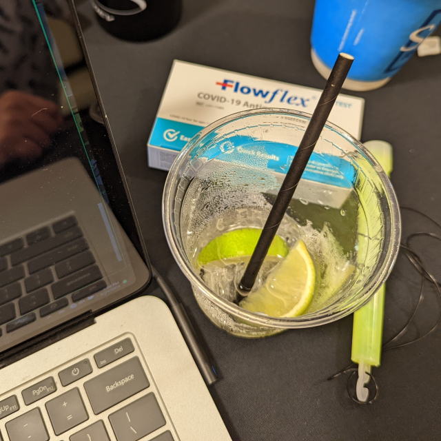 Looking down at a laptop, a Flowflex COVID home test, a glow stick that is still glowing, and an empty plastic cup with two lime wedges and a cocktail straw.